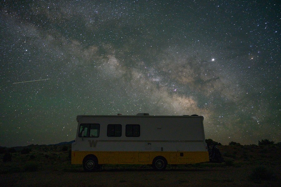 retro white and yellow RV underneath the Milky Way at night with shooting star in background